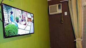 Una habitación con televisión instalada en la pared mostrando una escena, una ventana con cortinas y un equipo de aire acondicionado en la pared.