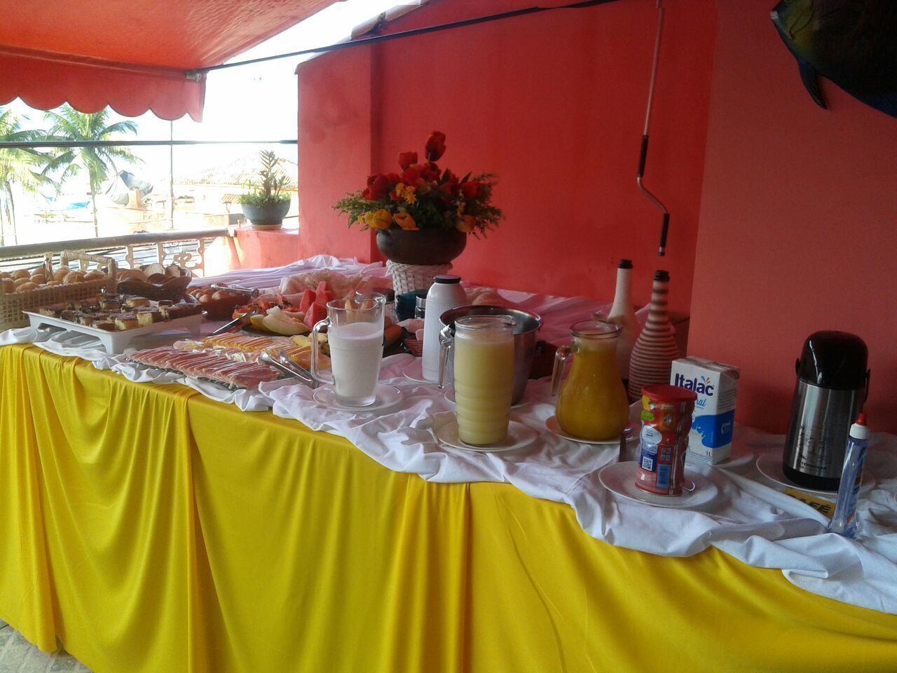 Uma mesa de buffet com diversos alimentos e bebidas, um vaso de flores e uma garrafa térmica.