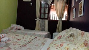 Una habitación con dos camas, ropa de cama floral, una ventana con cortinas, un aire acondicionado y cuadros en la pared.