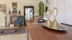 Un mostrador de recepción de madera con una bandeja que sostiene dos campanas de bronce, un sillón azul, una chimenea y un cuadro en la pared.