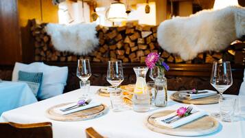 A dining table set with wine glasses, plates, and a pitcher of water, with a rustic wooden backdrop and a stone wall.