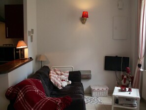 A living room with a dark sofa, a red blanket, a white teddy bear, a lamp, a flat-screen TV, and a small white table with a vase. - Dodo et tartines B&B (Bayeux)