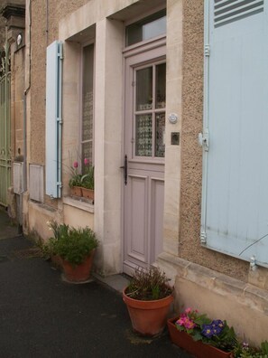 A building with a light-colored facade, a single door, and two windows with shutters. There are potted plants on the ground. - Dodo et tartines B&B (Bayeux)