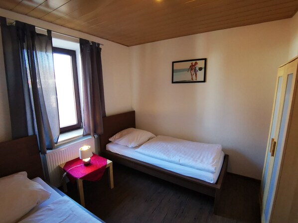 A small, neatly arranged bedroom with two single beds, a bedside table with a lamp, a window with curtains, and a framed picture on the wall. - Stadtdomizil (Augsburg)