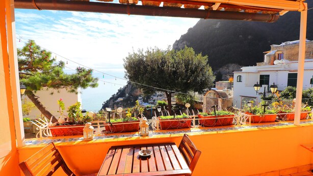 A terrace with a wooden table and chairs, potted plants, and a view of a coastal town with buildings and a mountain in the background. - Angolo di Paradiso (Positano)