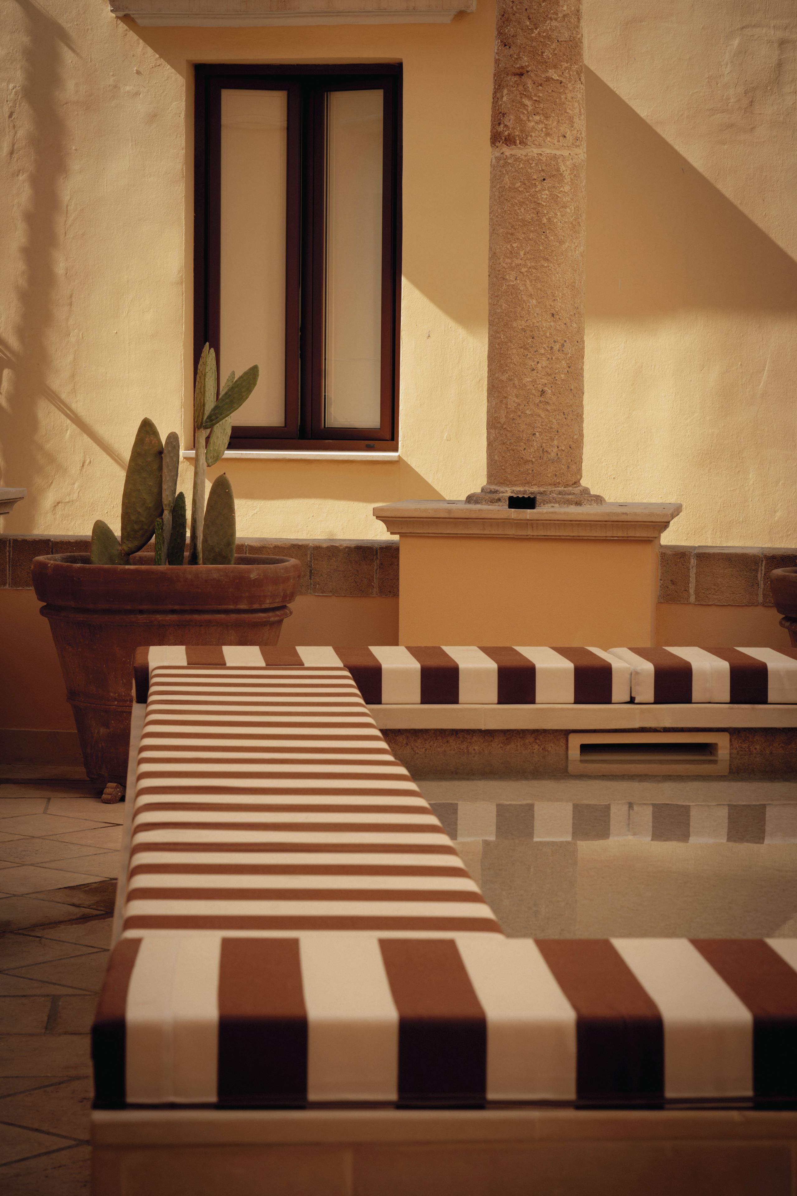 A sunlit outdoor seating area with striped cushions, a cactus plant, and a stone pillar.