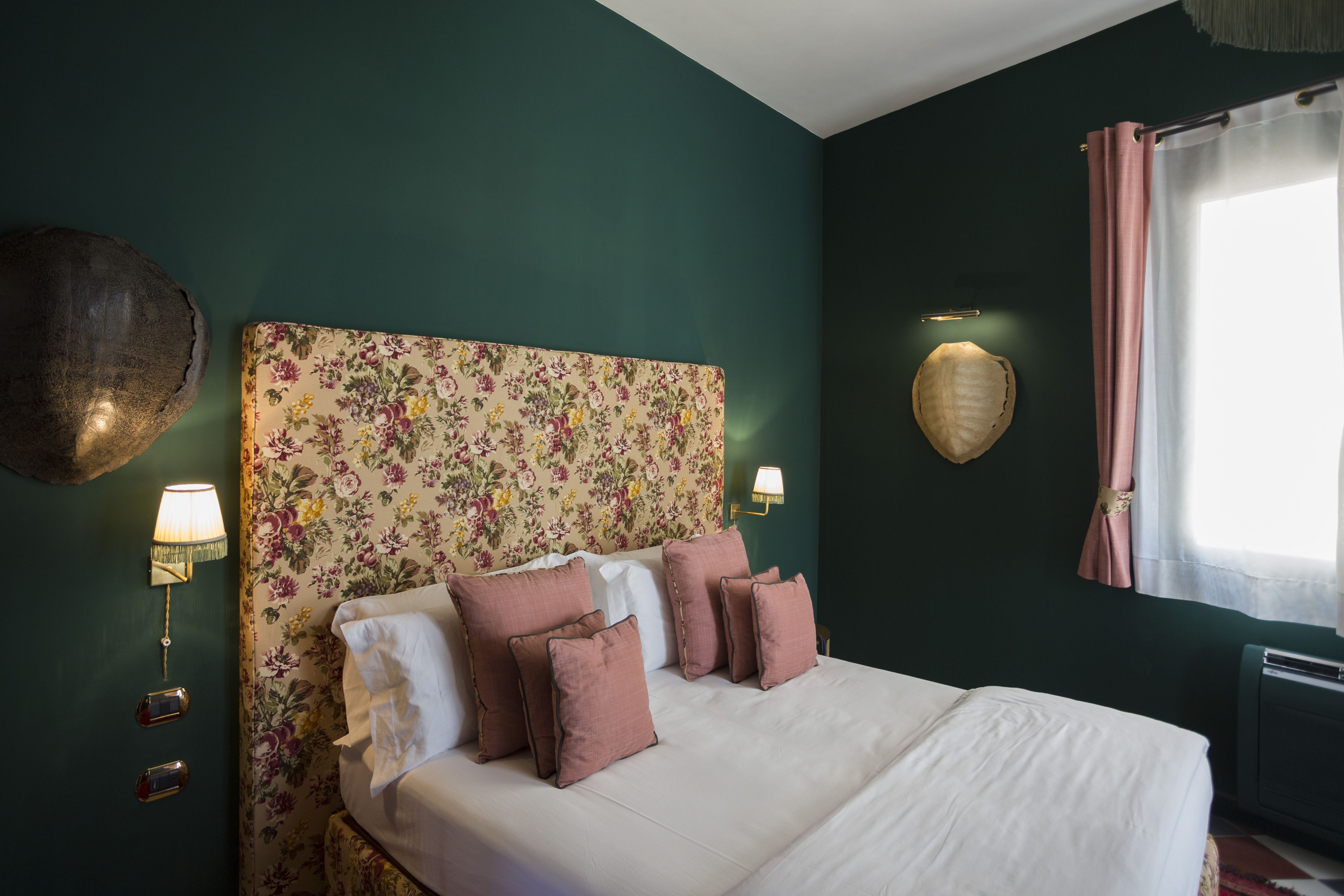 Un lit bien fait, avec une literie blanche et rose, une tête de lit à motifs floraux et des lampes fixées au mur.