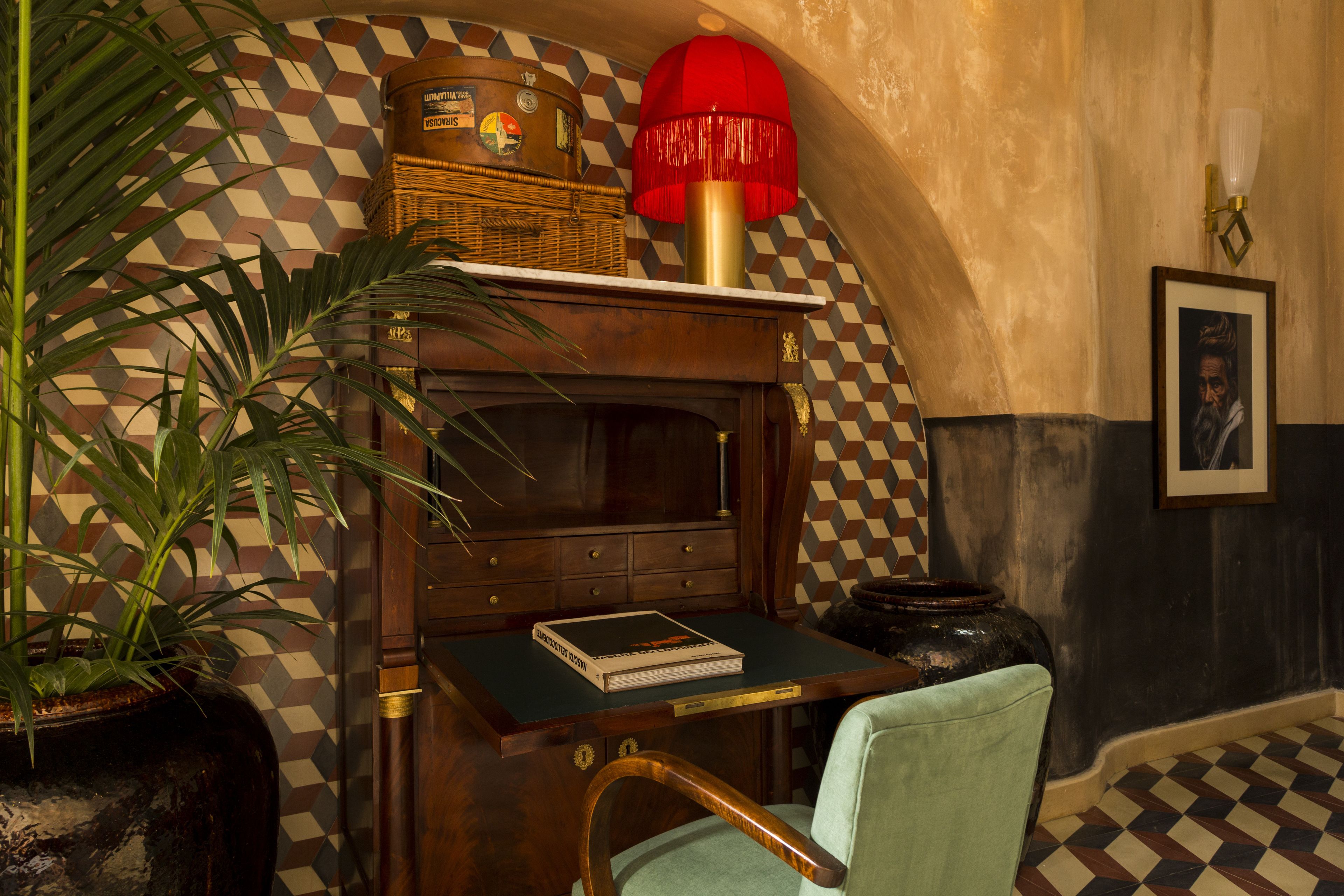 A room with a checkered floor, a wooden desk with a book, a red lamp, and a framed picture on the wall.