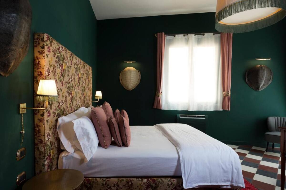 Un dormitorio con cama, mesita de noche, lámpara, reloj en la pared y ventana con cortinas.
