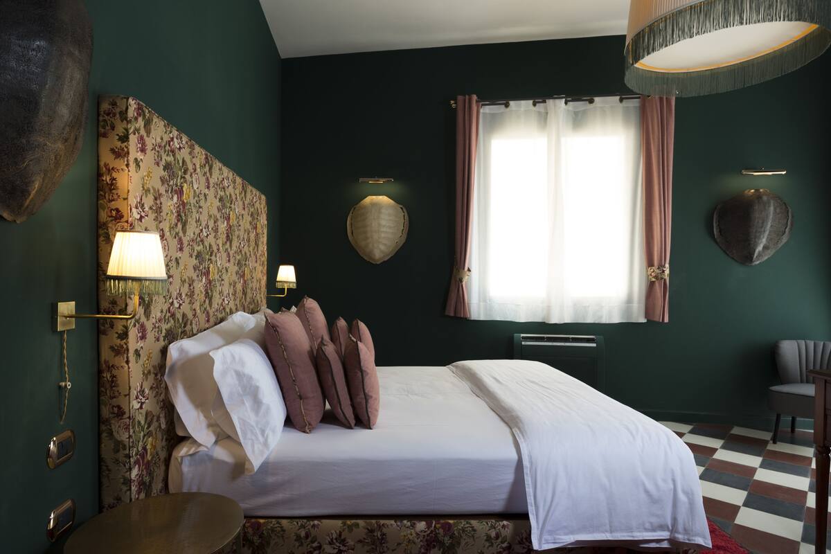 Una cama bien hecha con ropa de cama blanca y rosa, una mesa redonda y una silla en una habitación con paredes verde oscuro, cabecera con motivos florales y un ventanal con cortinas.