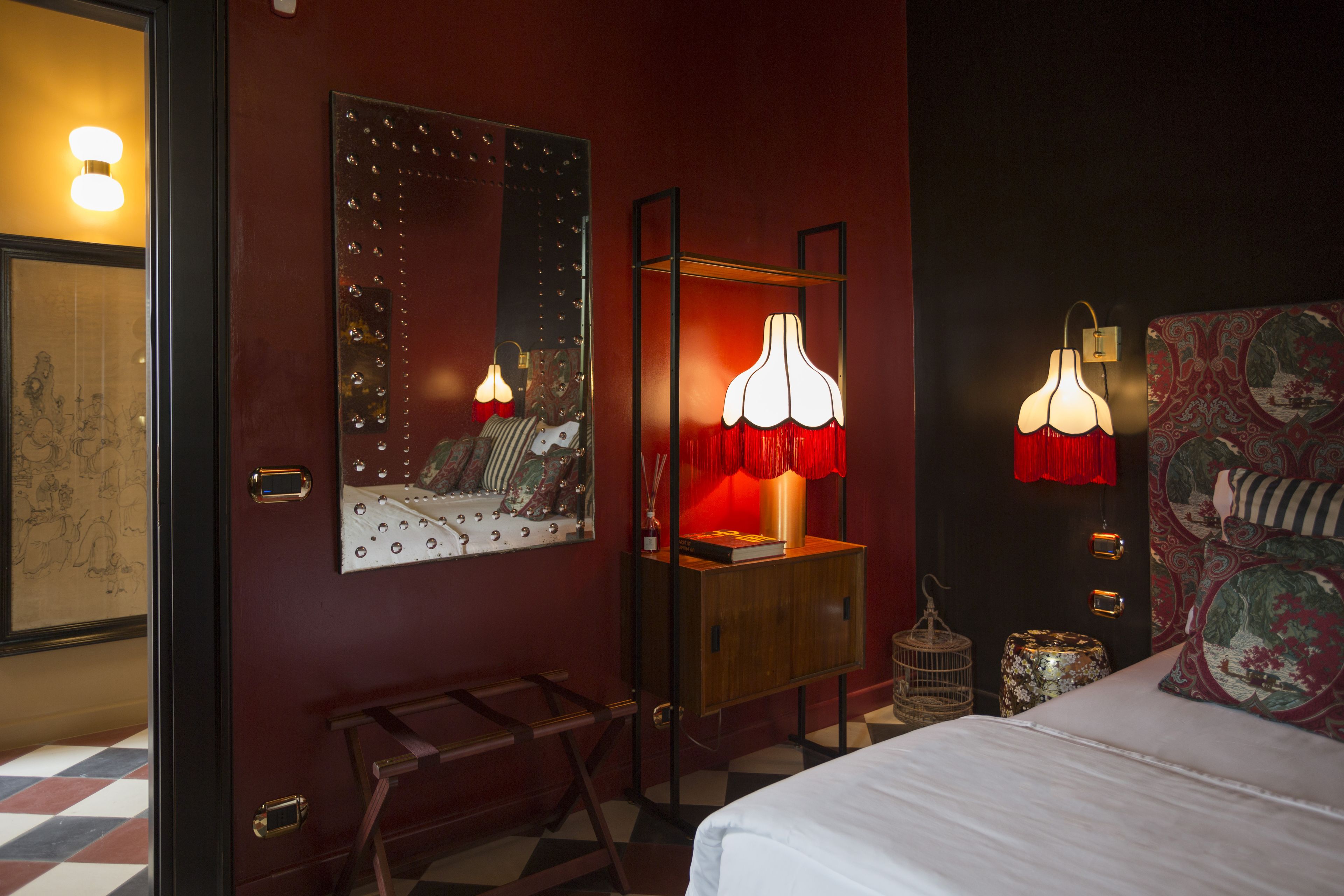 Una camera d'albergo con un letto grande, un comodino in legno, uno specchio e una lampada rossa.