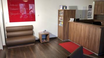 Un moderno vestíbulo de hotel con mostrador de recepción, una zona de estar con sofá, un pequeño mueble de madera con folletos y una pared roja con letrero de temperatura y restaurante.