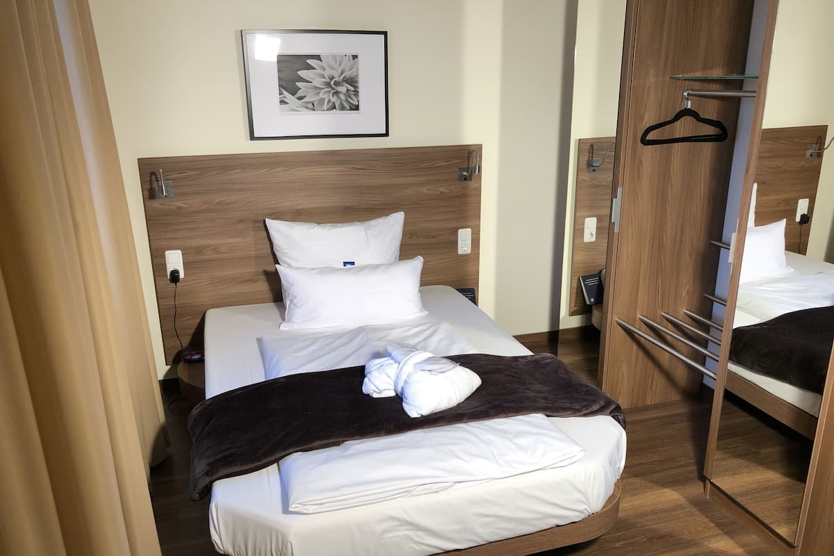 Una cama bien tendida con sábanas blancas, una colcha marrón oscuro y una toalla blanca. Encima de la cama hay un cuadro enmarcado en la pared y un cabecero de madera.