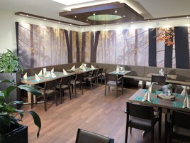 A restaurant with wooden tables and chairs, a large forest mural on the wall, and potted plants.