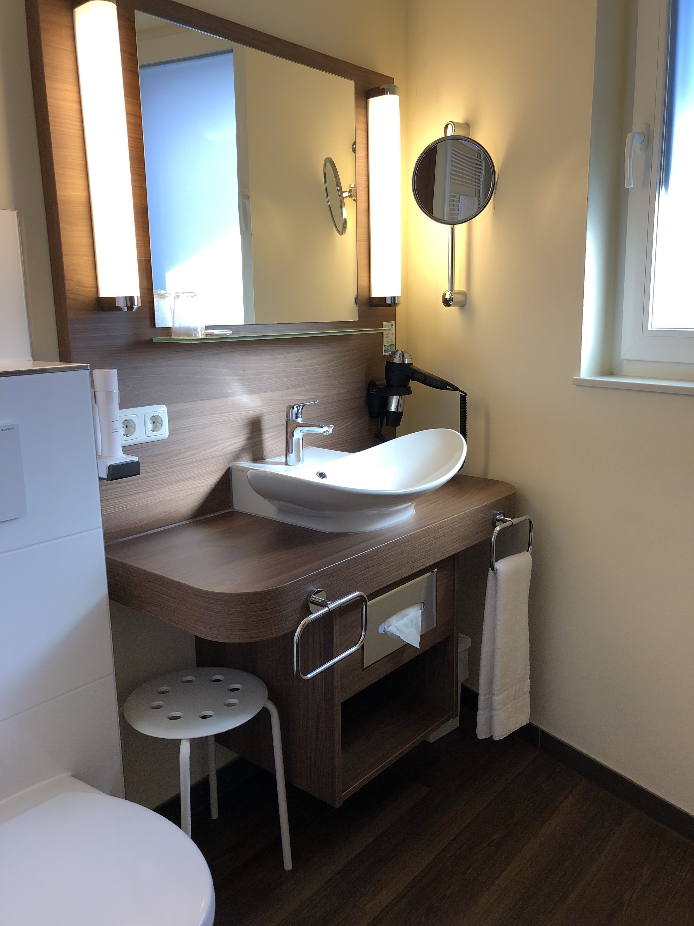 Un baño moderno con un lavamanos blanco, un espejo, un perchero y un taburete pequeño.