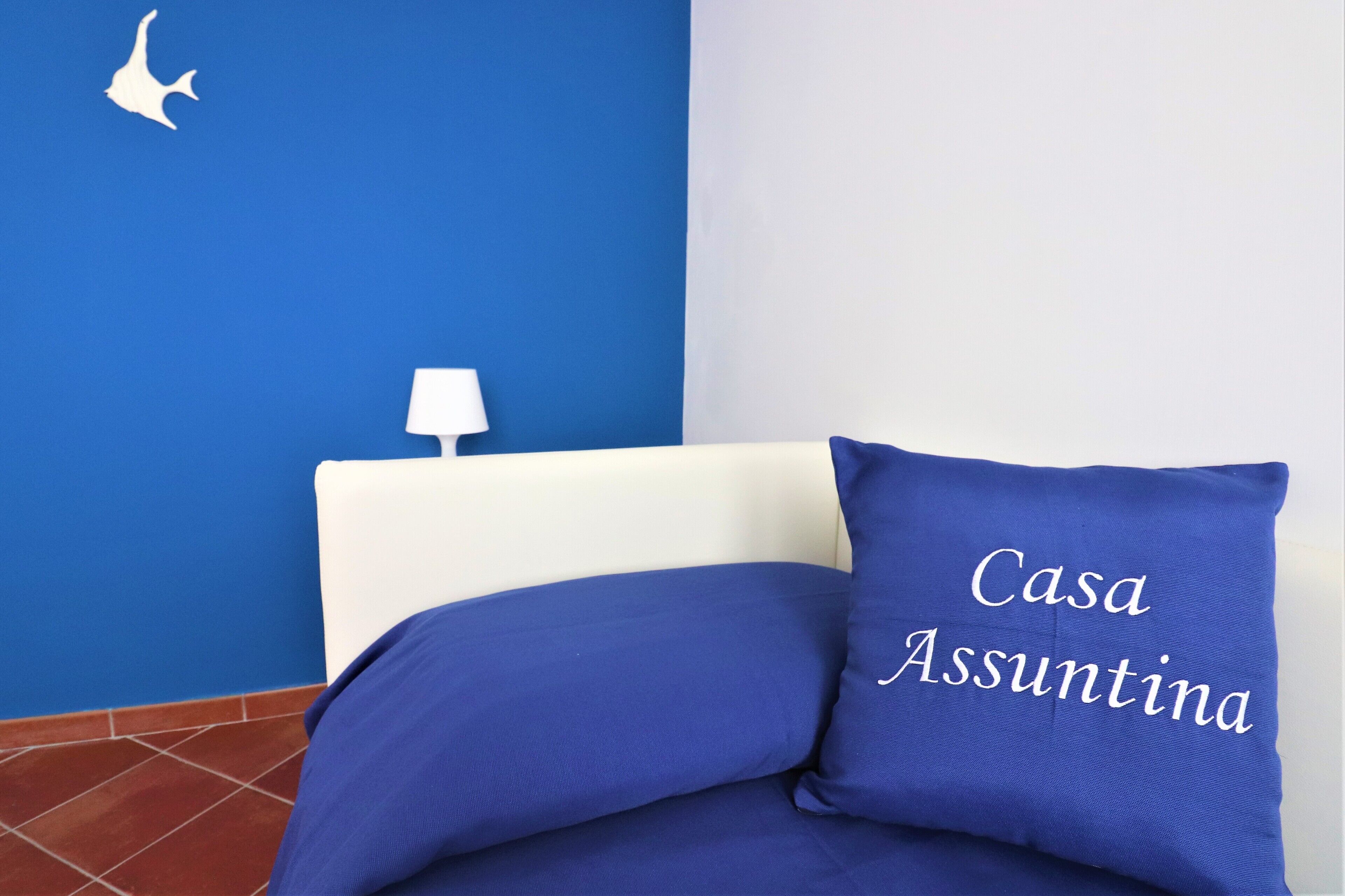 A bed with blue bedding and a pillow with "Casa Assuntina" text, a white lamp, and a blue wall with a white silhouette.