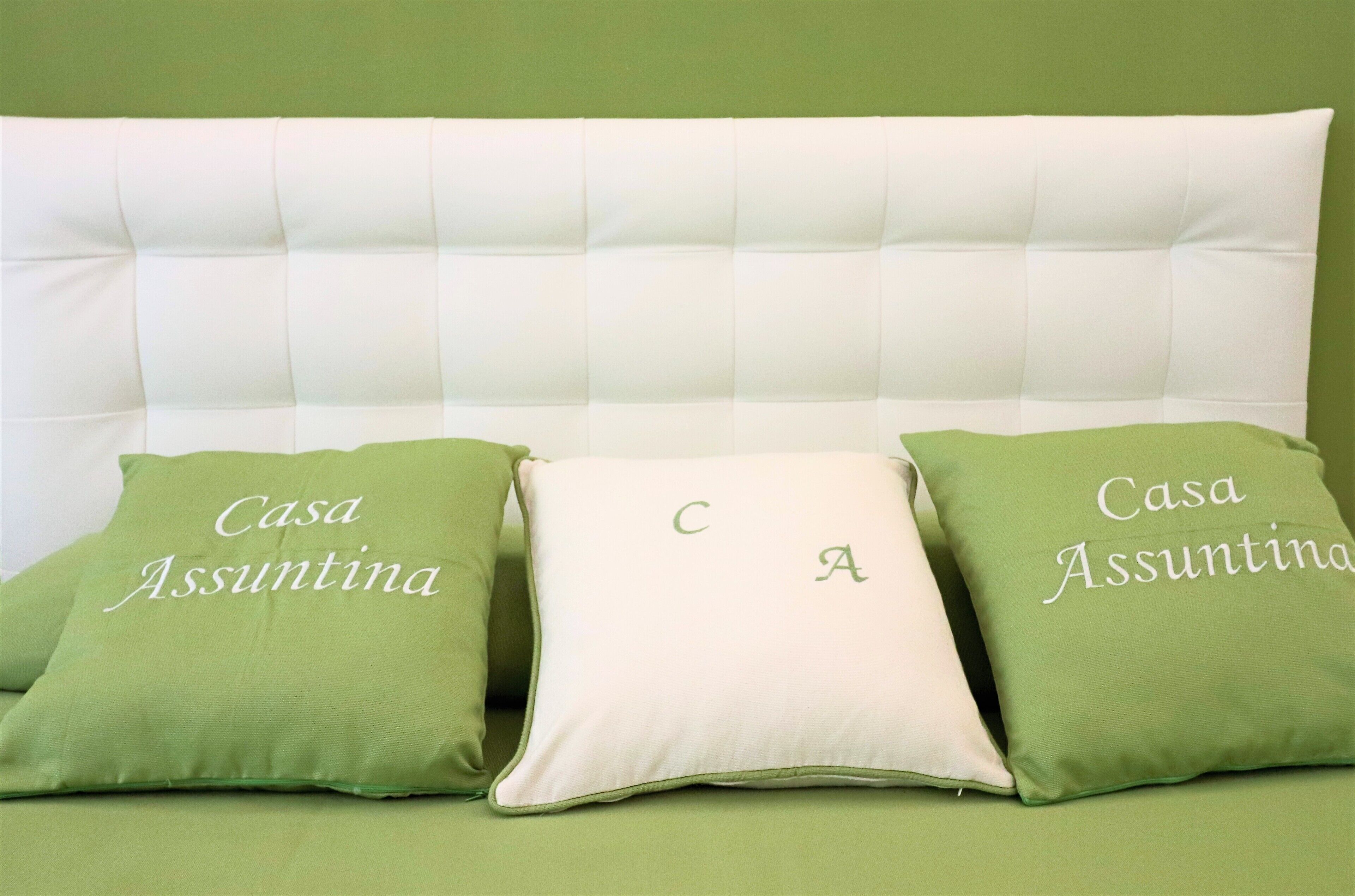 En säng med en vit sänggavel och tre kuddar, två gröna och en vit, med texten ”Casa Assuntina” på de gröna kuddarna.