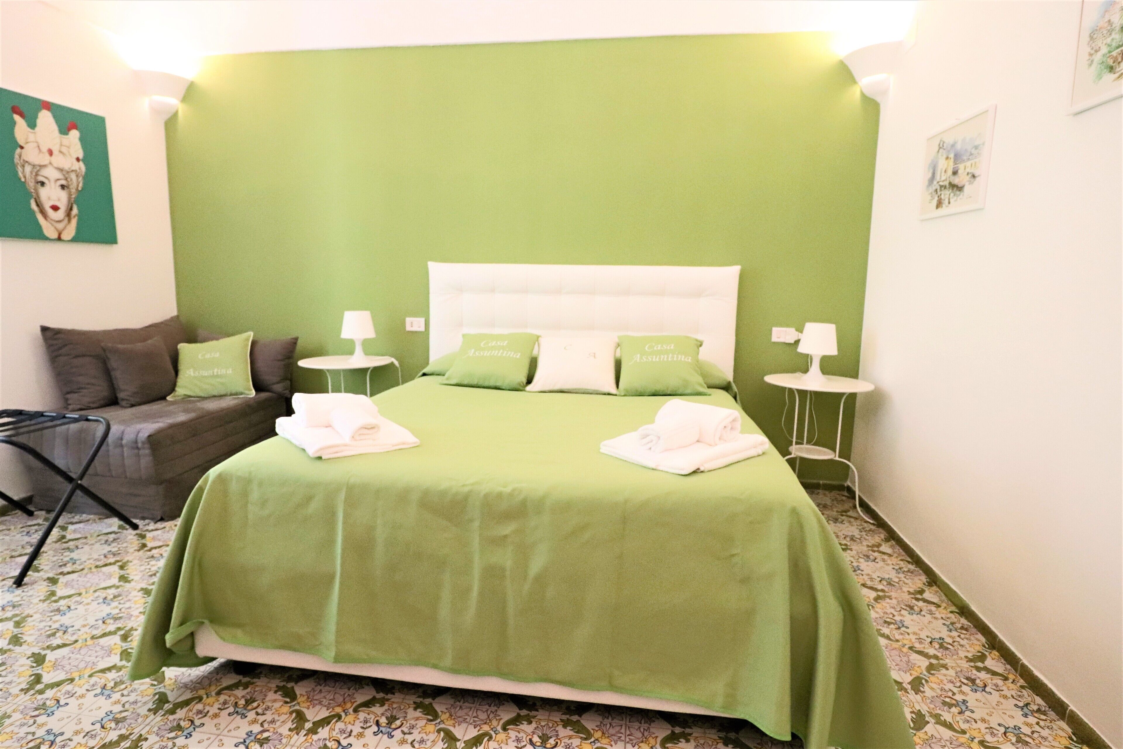 A neatly made bed with green bedding and pillows, a small bedside table with a lamp, and a patterned floor.