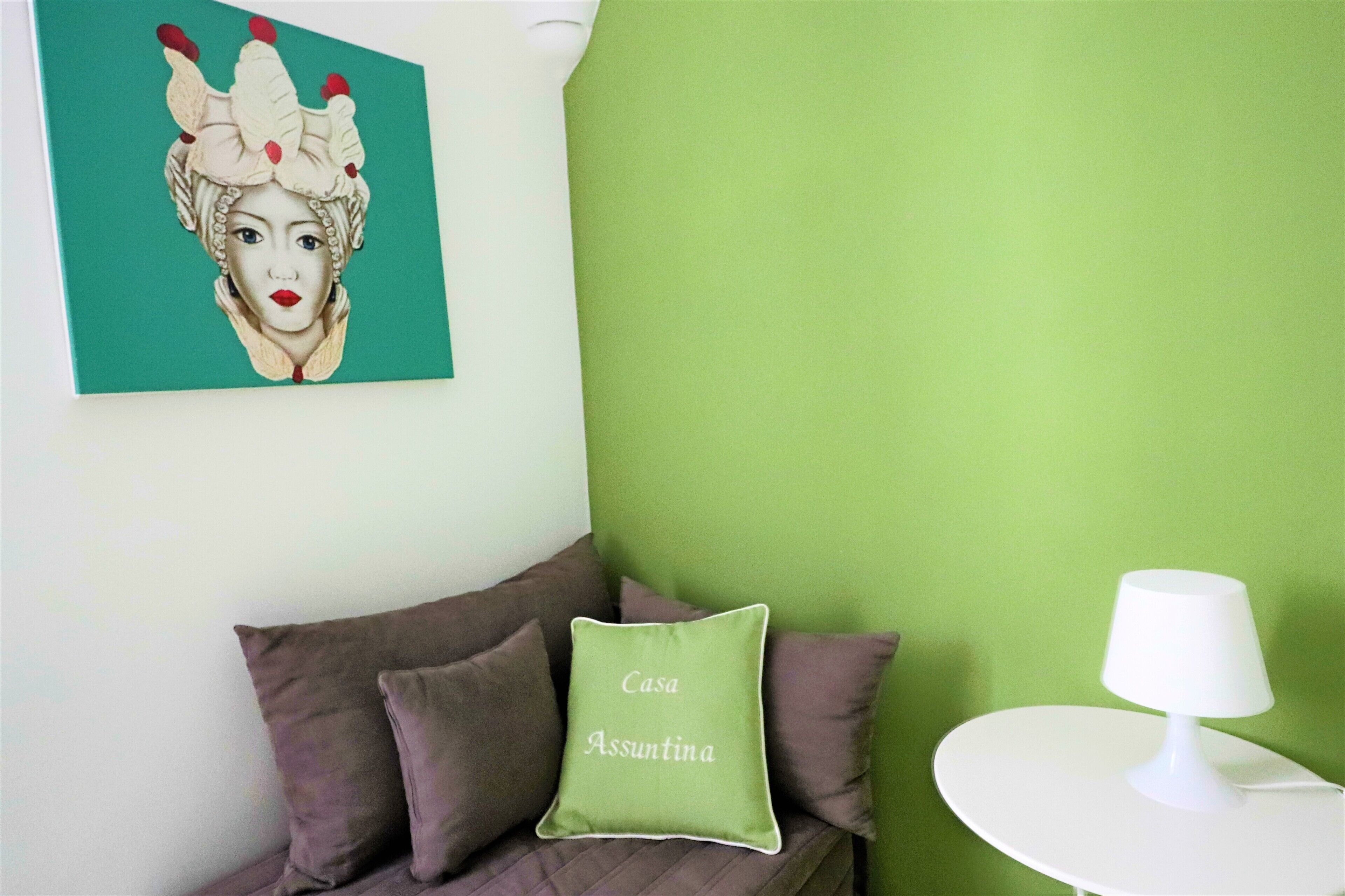 A room with a green wall, a white round table with a white lamp, and a couch with brown cushions and a green pillow labeled "Casa Assuntina".