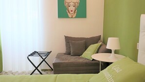 Een kamer met een bed, een klein tafeltje met een lamp, een aan de muur hangend schilderij en een rek voor wasgoed.