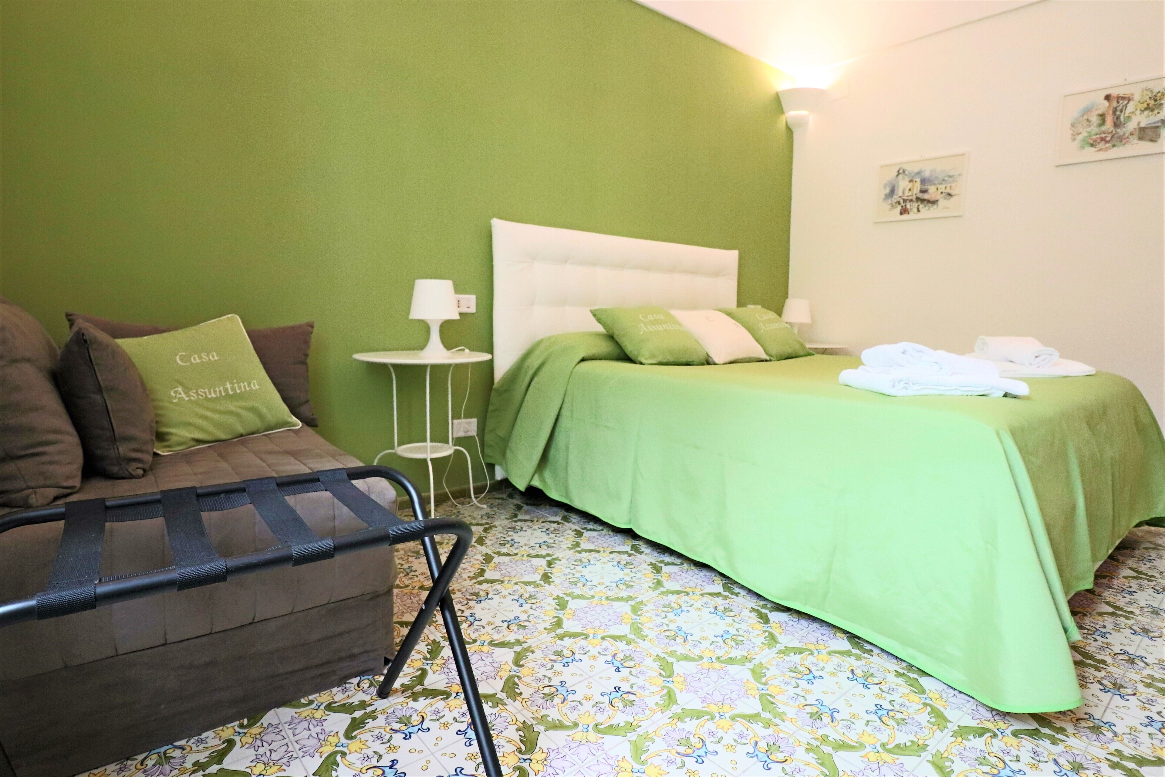 A bedroom with a green bedspread, a brown sofa with a cushion, a small bedside table with a lamp, and a patterned floor.