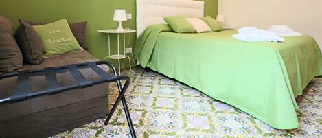 A bedroom with a green bedspread, a brown sofa with a cushion, a small bedside table with a lamp, and a patterned floor.
