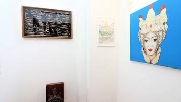 A room with a framed artwork of scissors, a framed painting of a building, and a large painting of a woman with a crown.