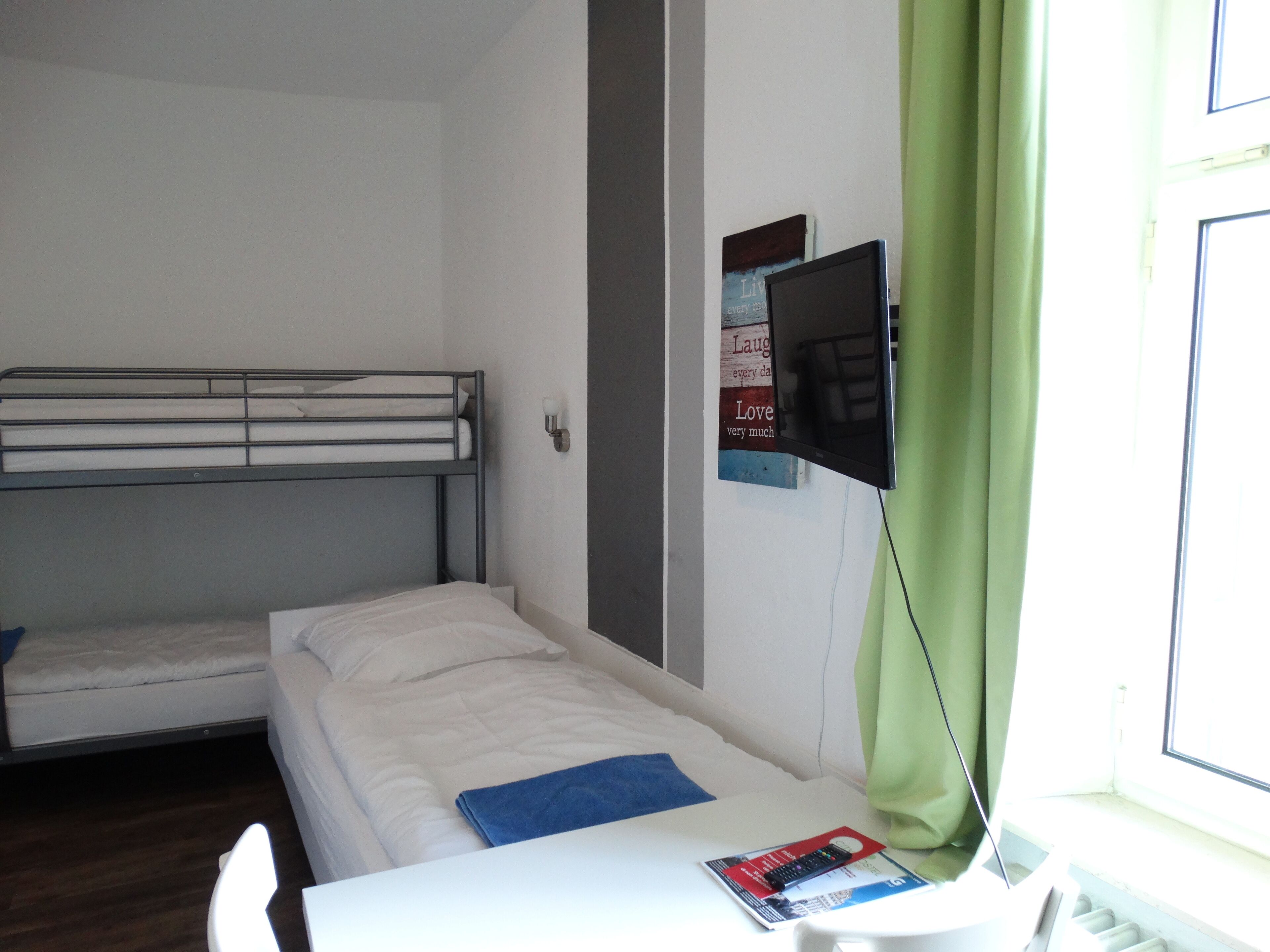 Ein Zimmer mit Etagenbetten, einem normalen Bett, einem an der Wand befestigten Fernseher und einem Fenster mit grünen Vorhängen.