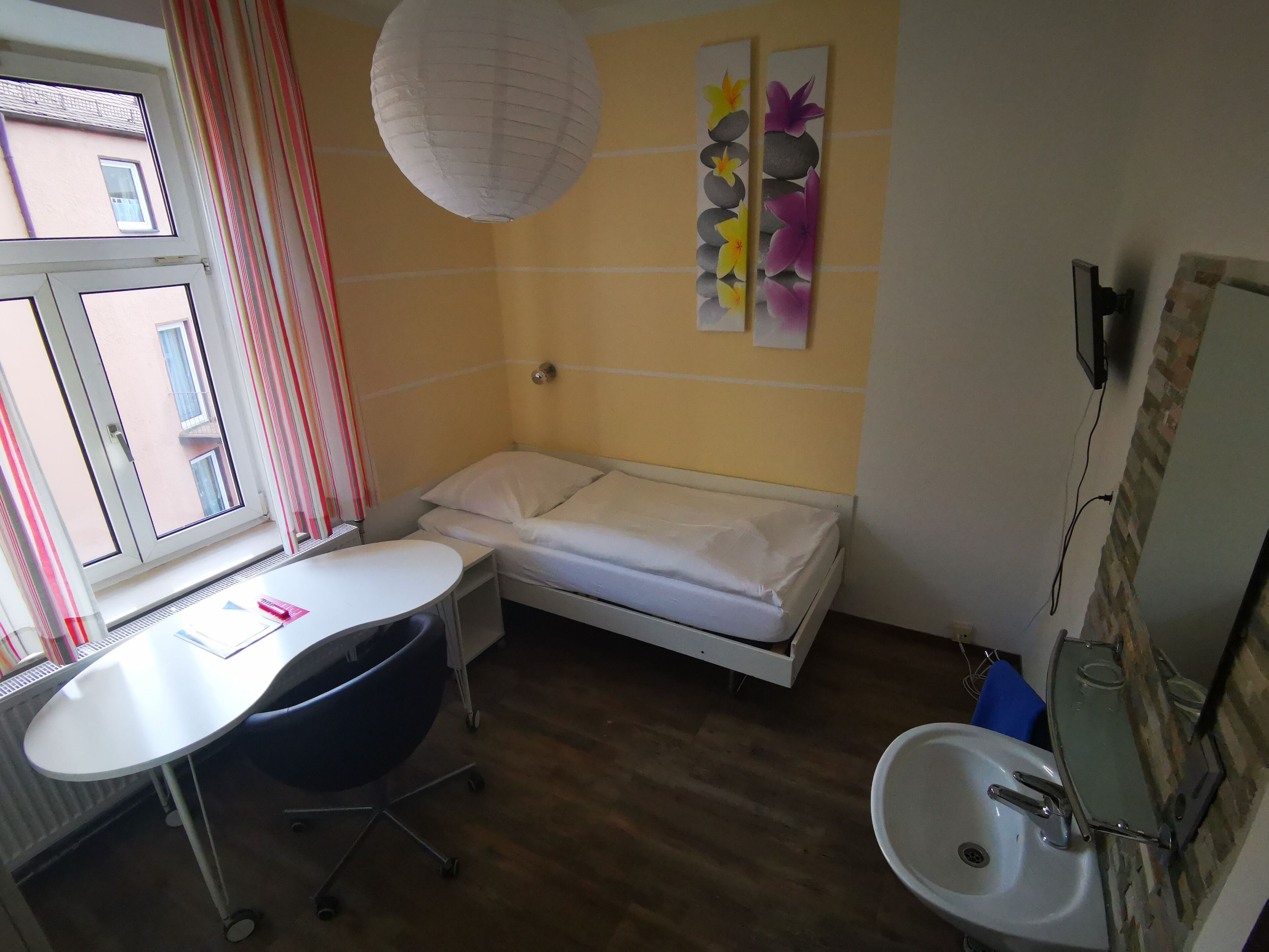 Quarto pequeno e compacto com uma cama de solteiro, uma escrivaninha e uma cadeira. Possui uma janela com cortinas, uma pia fixa na parede e um espelho.