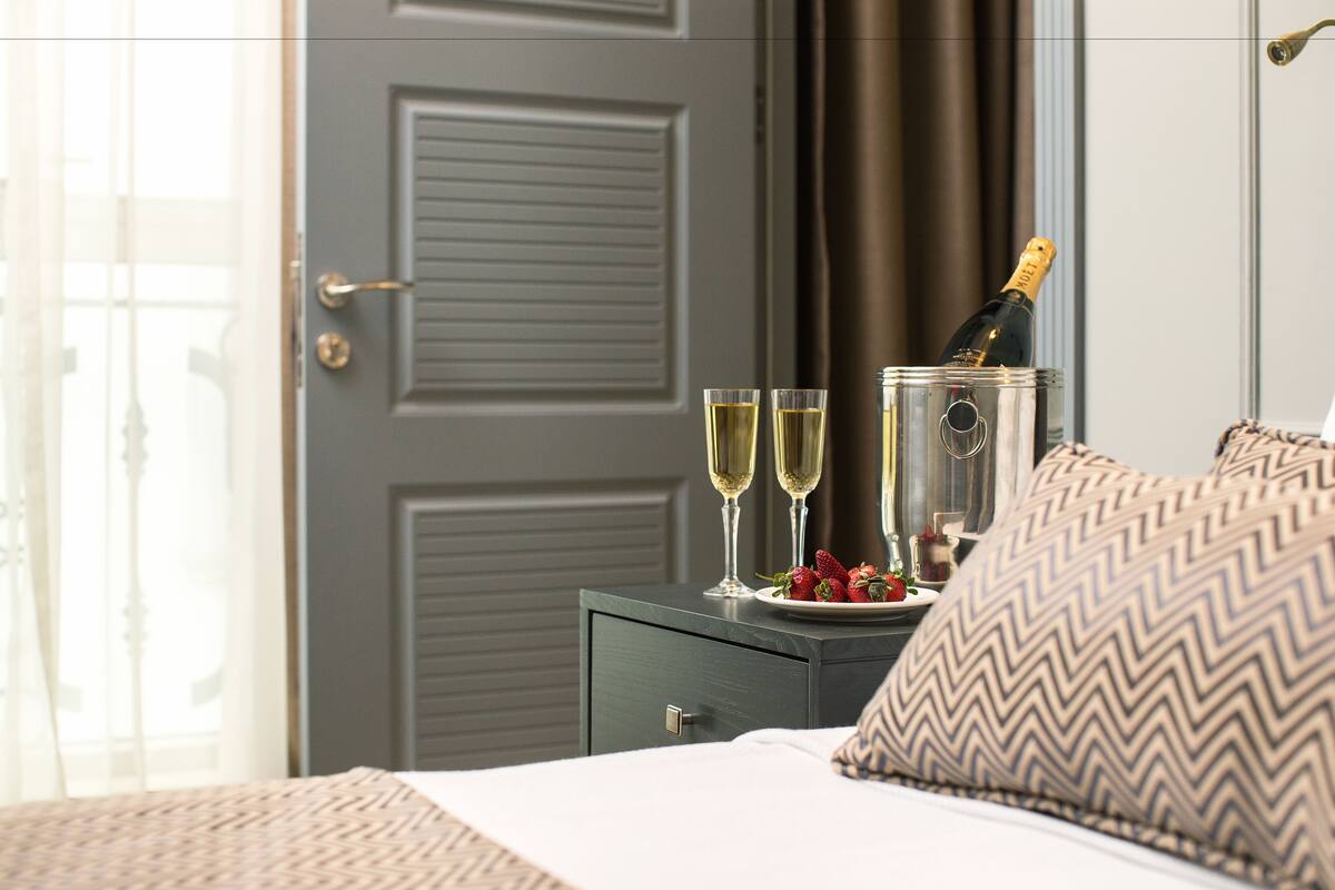 A neatly made bed with a pillow, a bedside table with a bottle of champagne, two glasses of champagne, and a plate of strawberries.