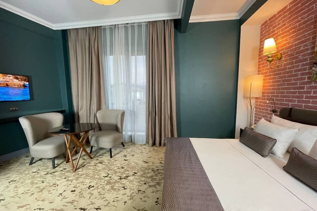 A hotel room with a bed, two armchairs, a small table, a TV, and a brick wall with a lamp.