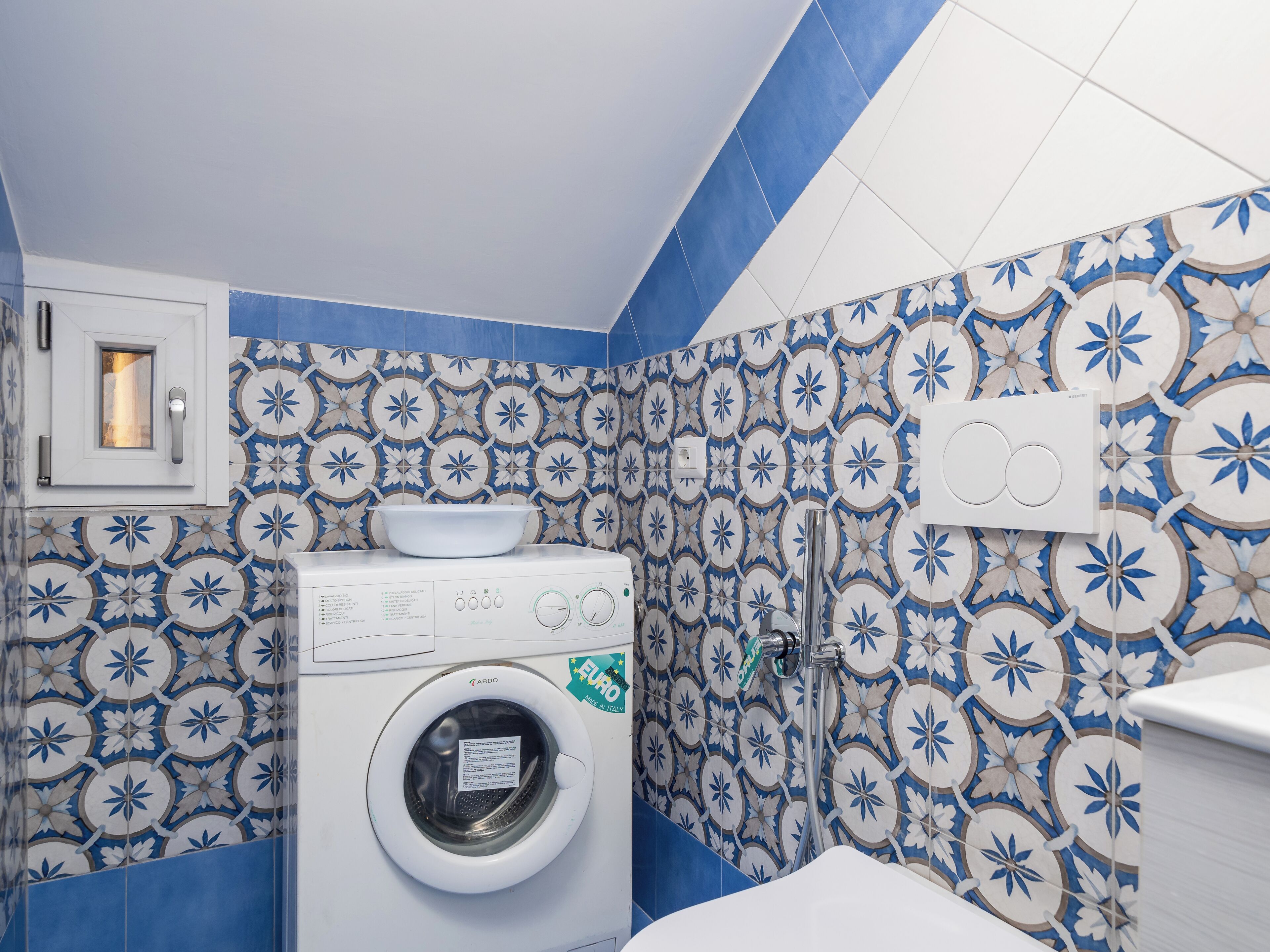 A compact laundry room with a washing machine, a sink, and a shower. The walls are tiled with a blue and white floral pattern.
