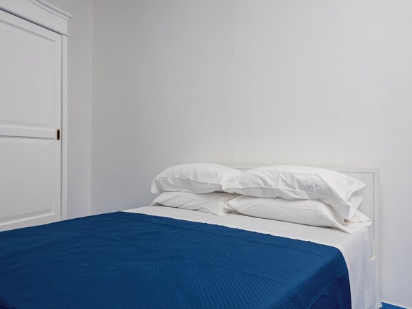 A bed with white bedding and a blue blanket, a white door, and a plain wall. - De Vivo Realty - Casa Alessandra. (Positano)