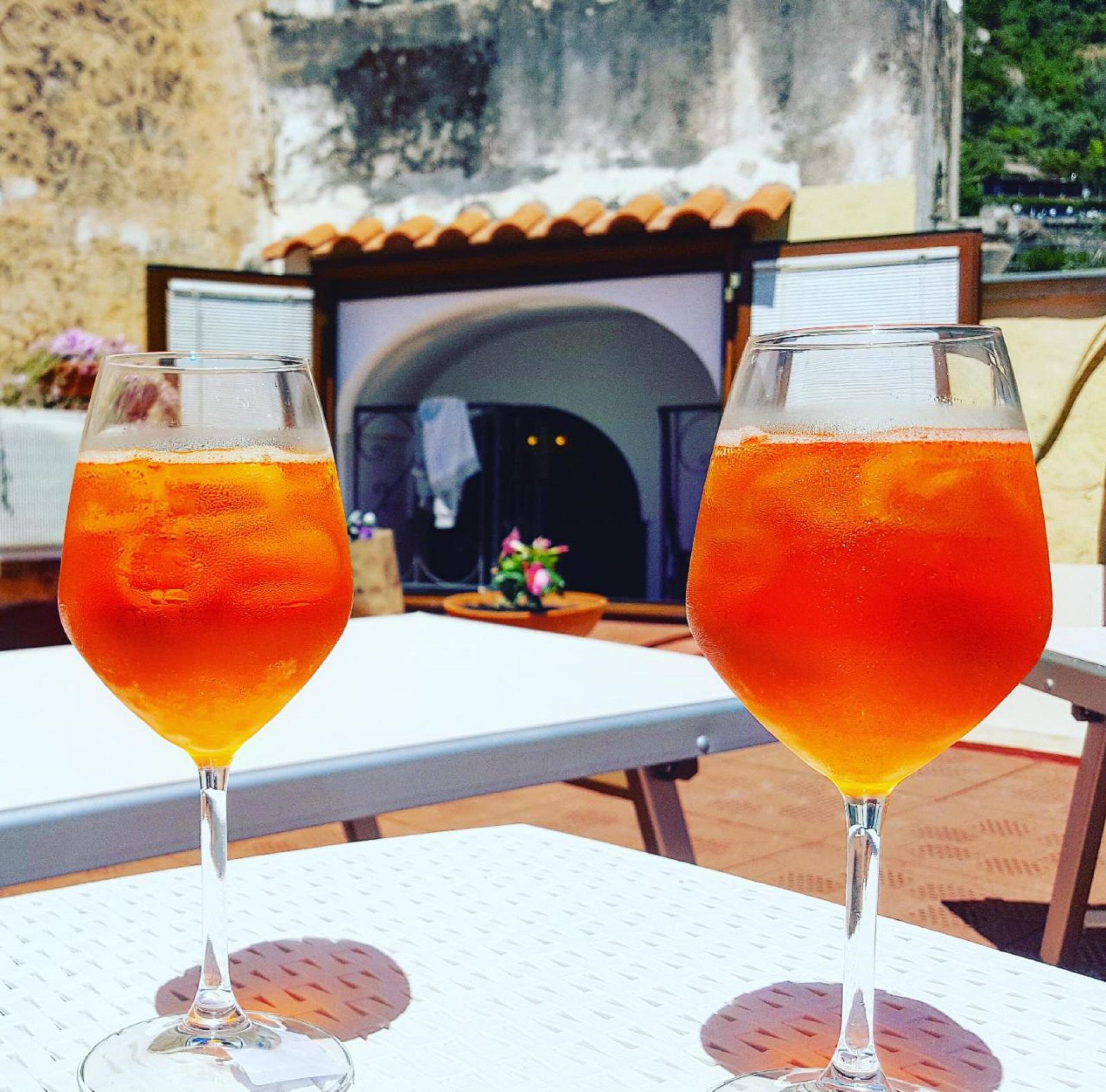 Two glasses of orange beverage on a table with a view of a building and a garden.