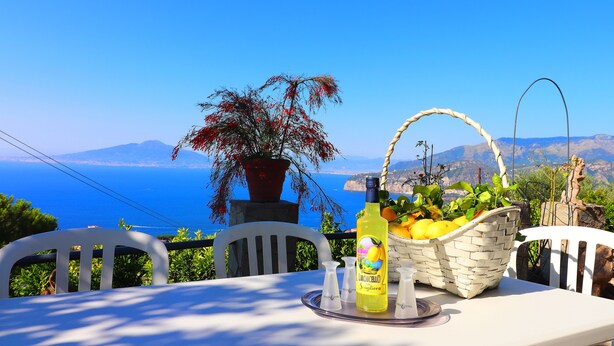Un tavolo con una bottiglia di Limoncello e un cesto di frutta, con vista su un paesaggio costiero panoramico.