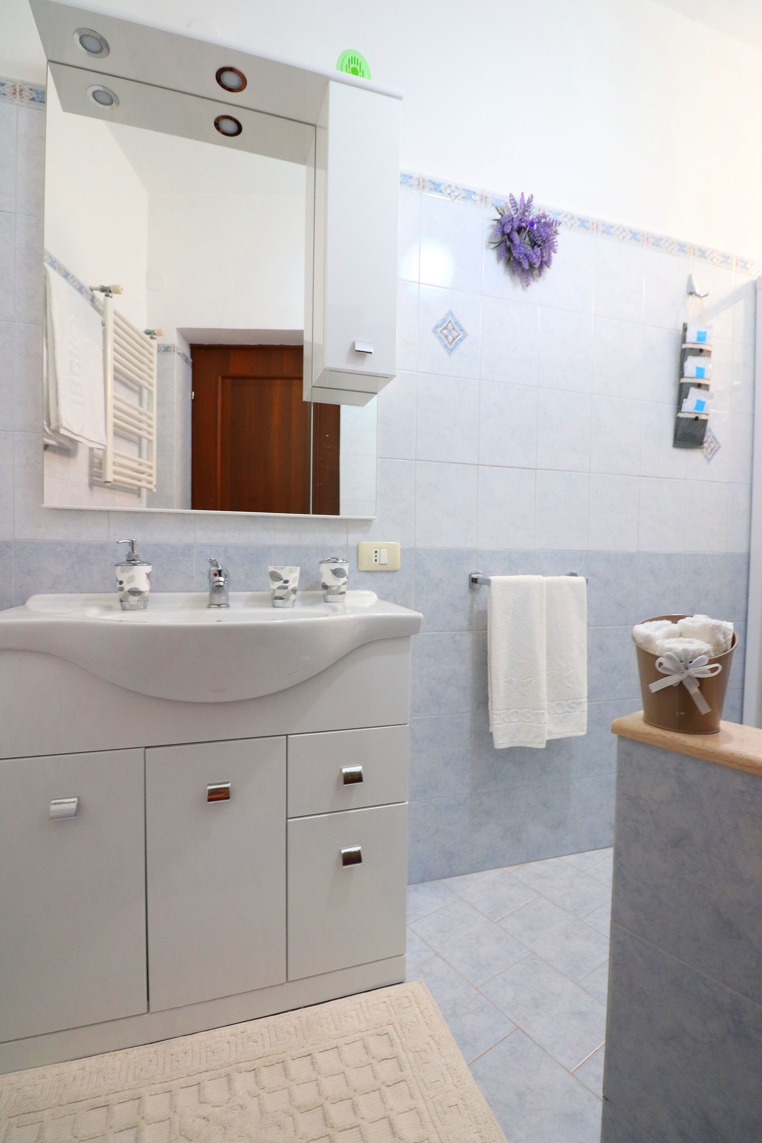 Um banheiro com pia branca, três torneiras, um espelho, porta-toalhas e uma flor roxa decorativa na parede.