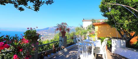 Une terrasse avec des chaises blanches et une table, offrant une vue panoramique sur la mer et les montagnes.