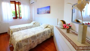 Una camera d'albergo con due letti, una finestra con tende, un quadro che raffigura uno skyline cittadino, un vaso con fiori e uno specchio.