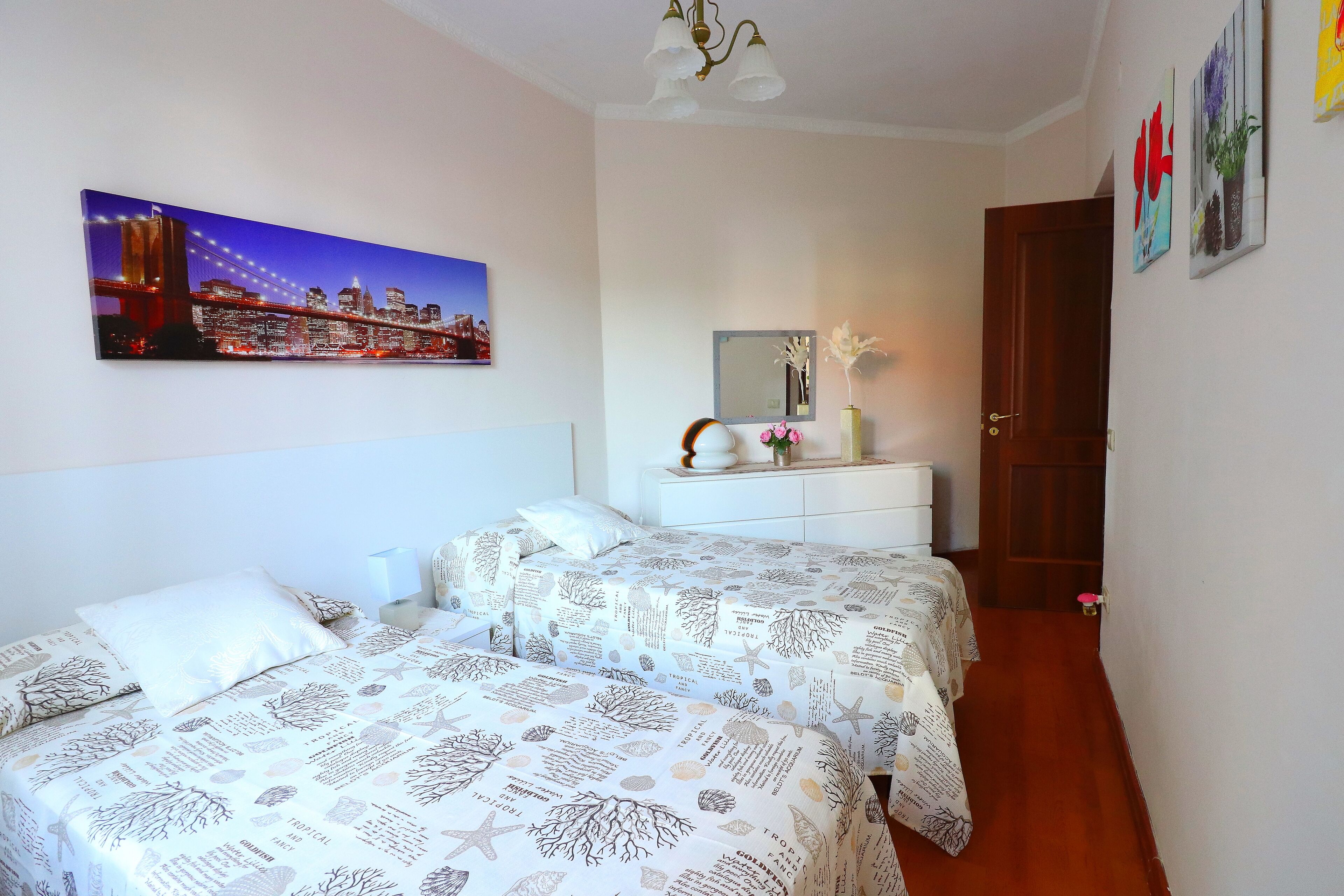 Quarto com duas camas, mesa de cabeceira, cômoda, espelho e pintura de paisagem urbana.