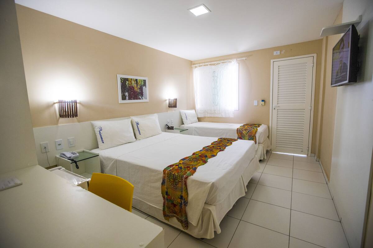 A hotel room with two beds, a desk, a chair, a window with curtains, a TV, and a wall-mounted lamp.
