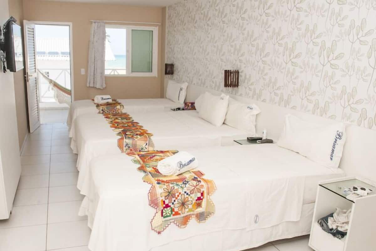A hotel room with a long bed, a small bedside table, a nightstand, and a view of the sea through a window.