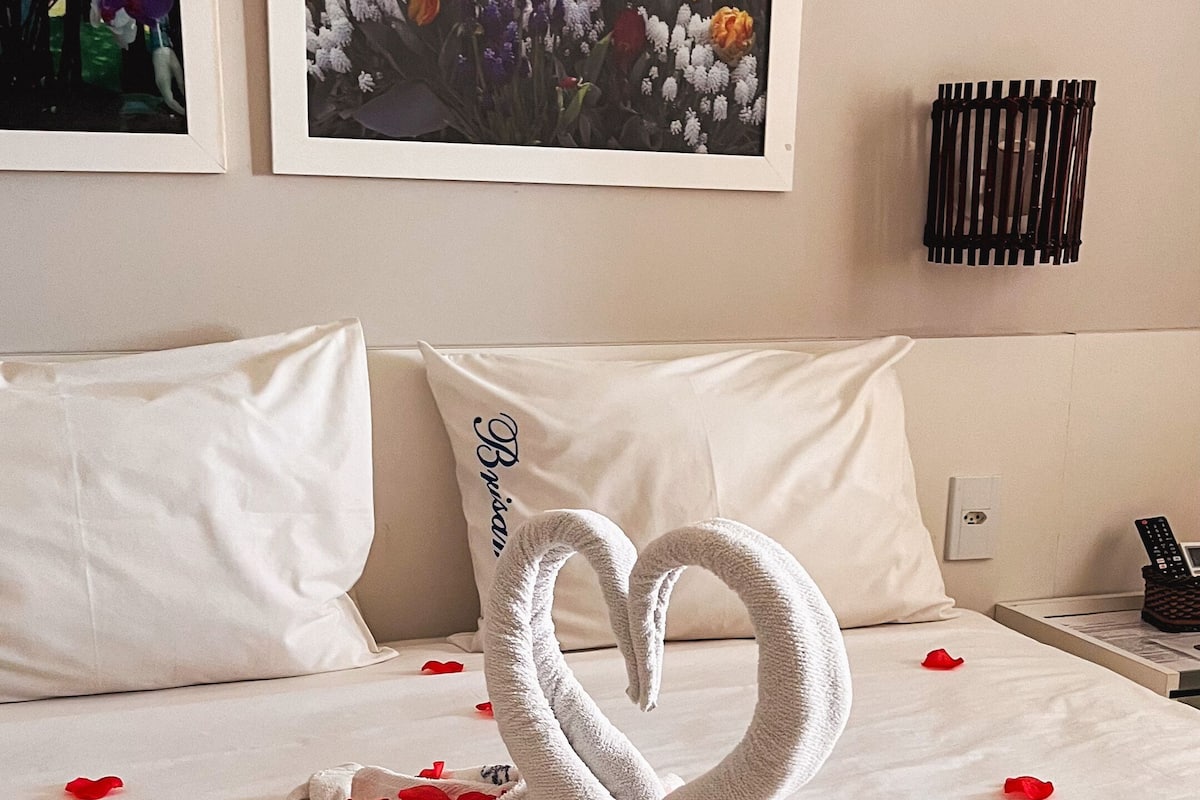 A hotel room with a bed, white pillows, a heart-shaped towel arrangement, and two framed floral pictures on the wall.