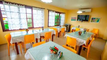 A dining area with multiple tables and chairs, large windows with lace curtains, and a television on the wall.