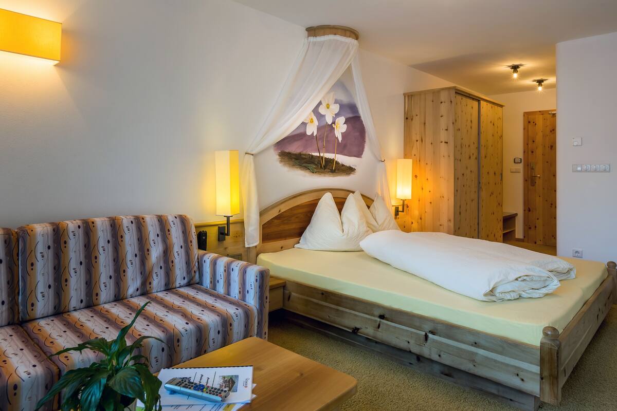 A hotel room with a bed, a sofa, a wooden wardrobe, and a wall-mounted artwork.
