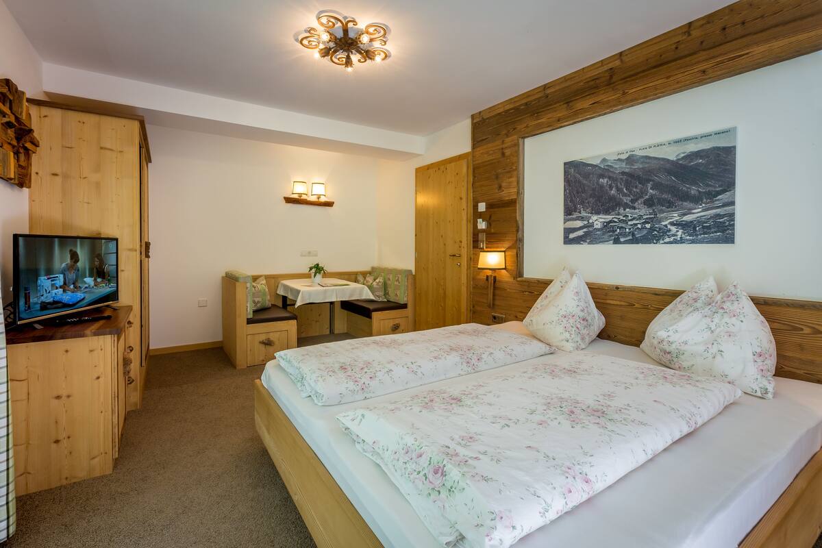 A bedroom with a wooden headboard, a bed with floral bedding, a television on a wooden cabinet, a desk with a lamp, and a framed picture on the wall.