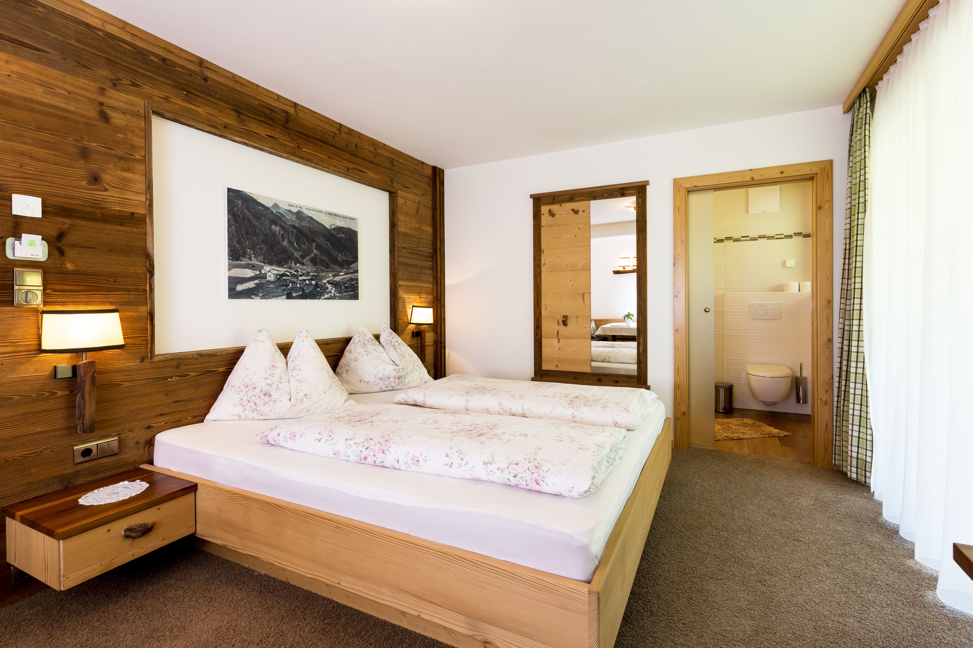 Un dormitorio con estructura de madera, una cama grande, una mesita de noche, una lámpara de pared y un cuadro enmarcado con un paisaje montañoso.