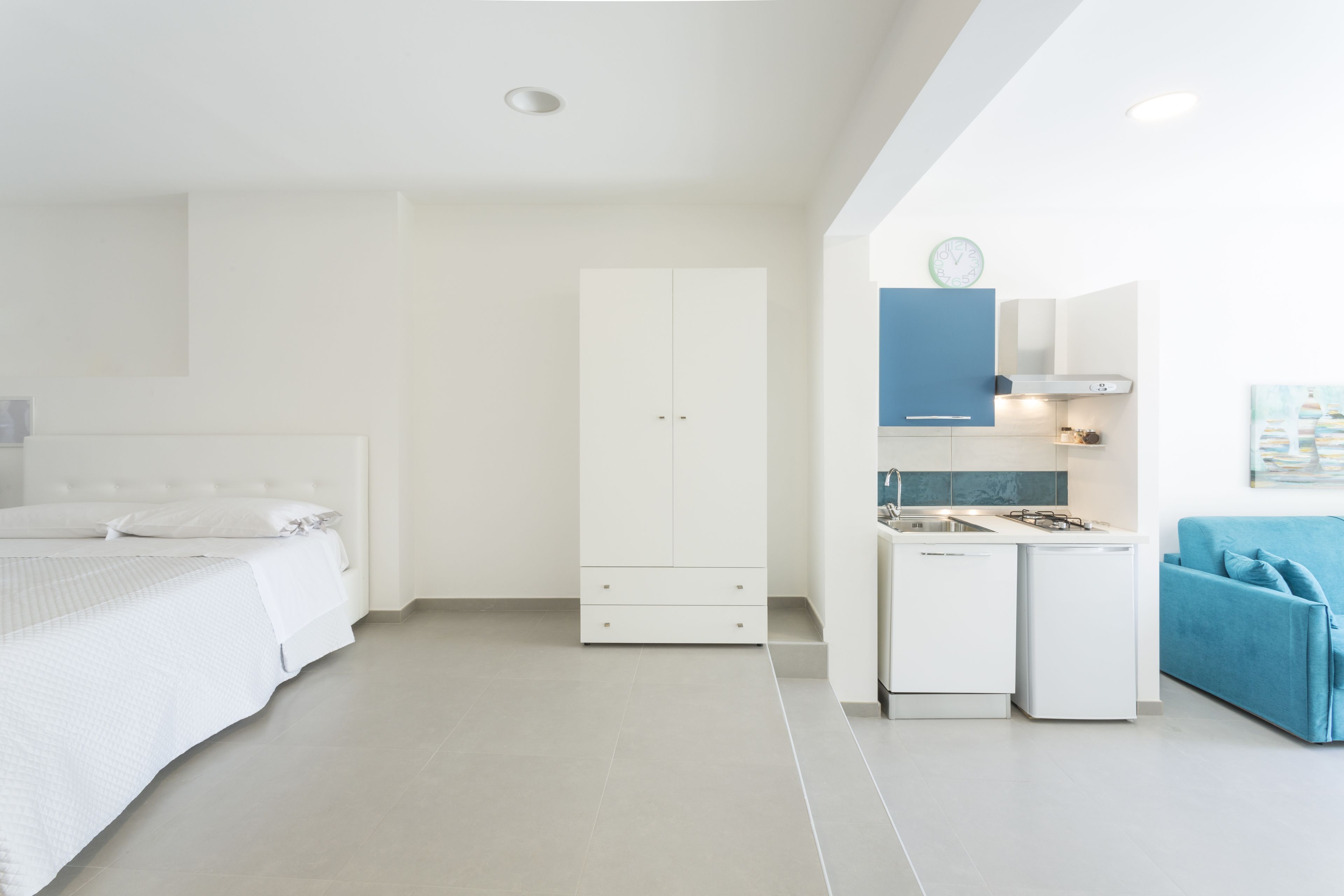 Un dormitorio moderno con cama blanca, armario, una pequeña kitchenette y sofá azul.