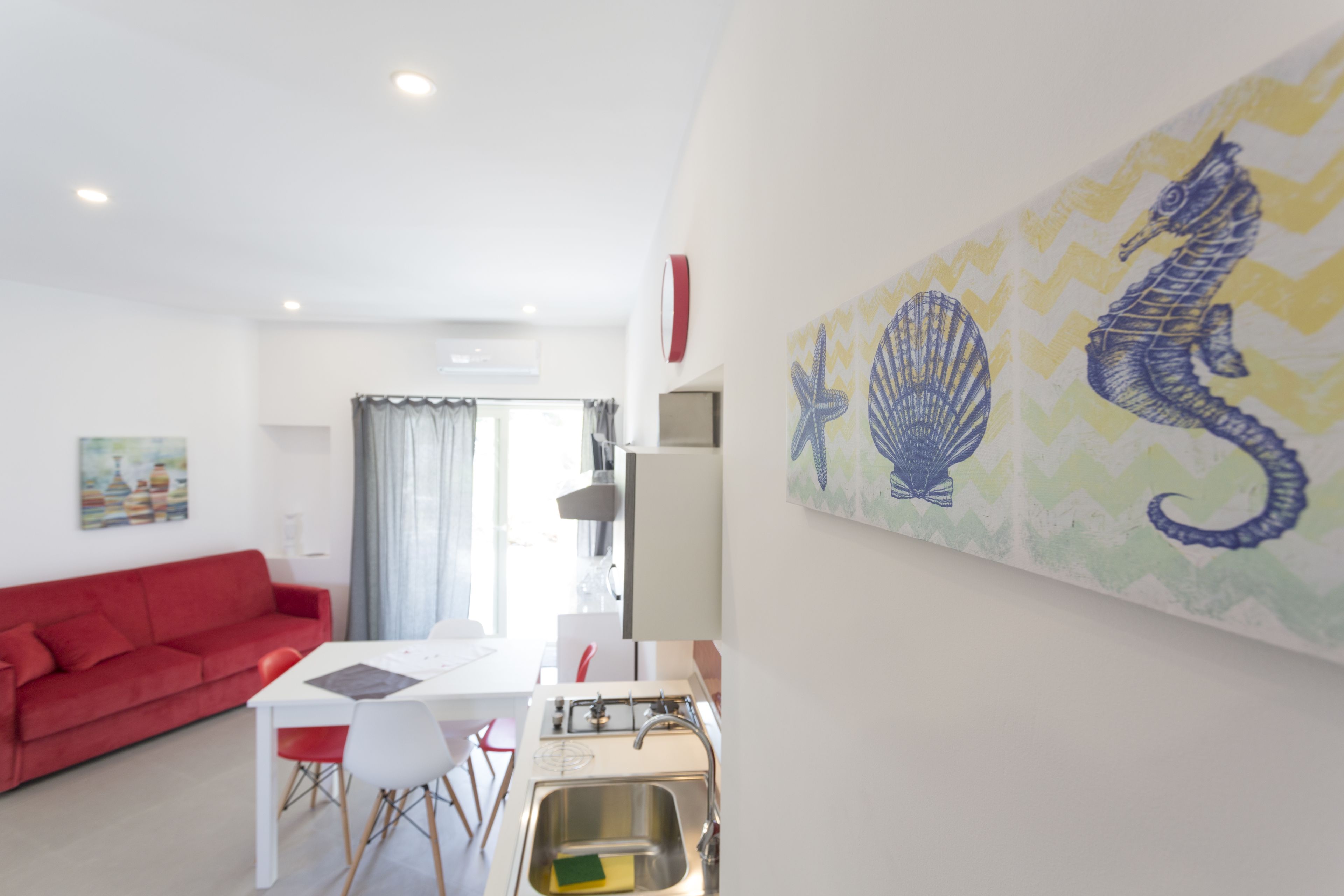 A modern living room with a red sofa, a dining table with chairs, a kitchen area with a sink, and a wall with a seahorse painting.