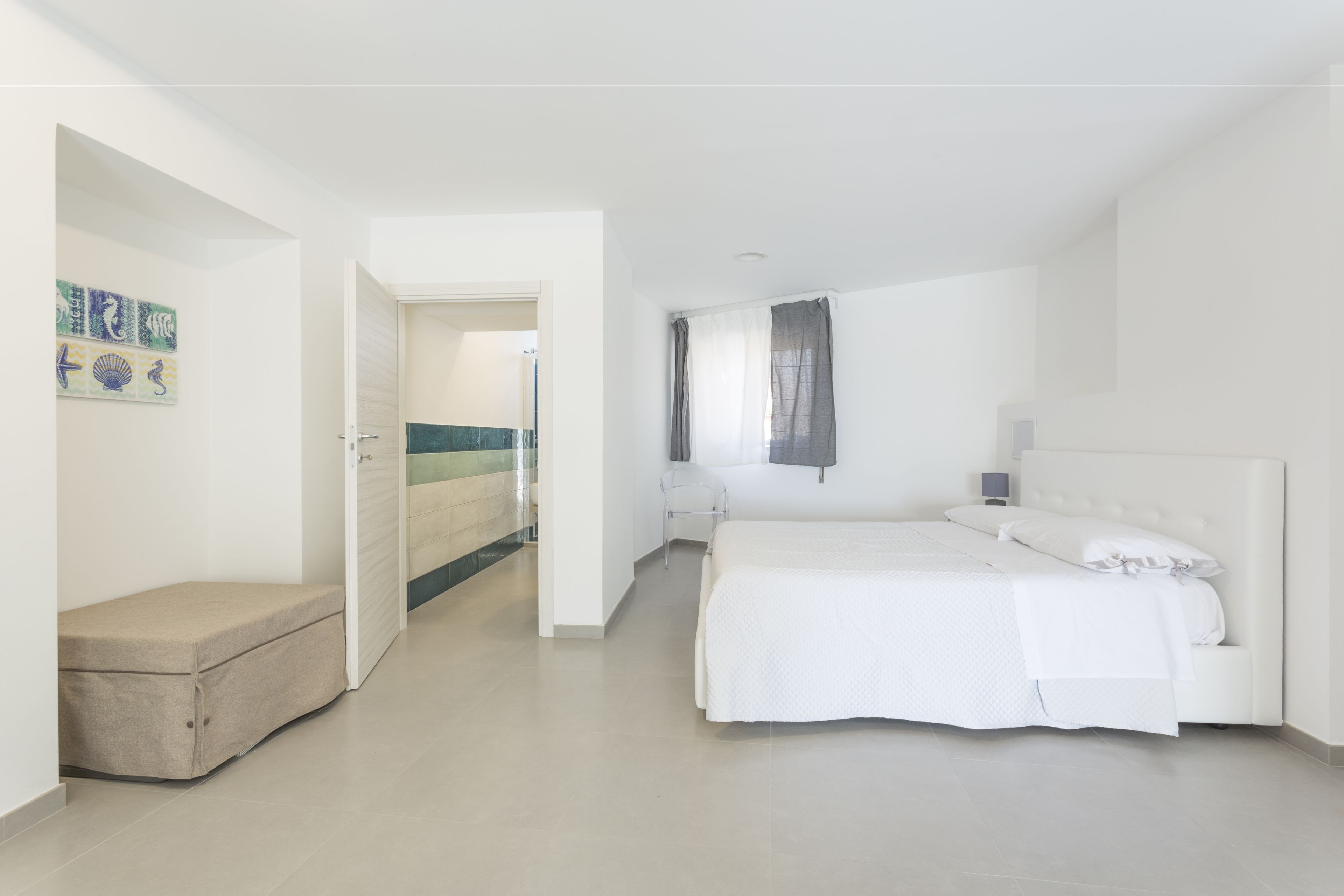 A modern bedroom with a large bed, a bedside table, and a chair. There is a door leading to another room and a small bench in the corner.