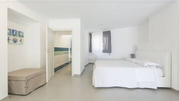 A modern bedroom with a large bed, a bedside table, and a chair. There is a door leading to another room and a small bench in the corner.